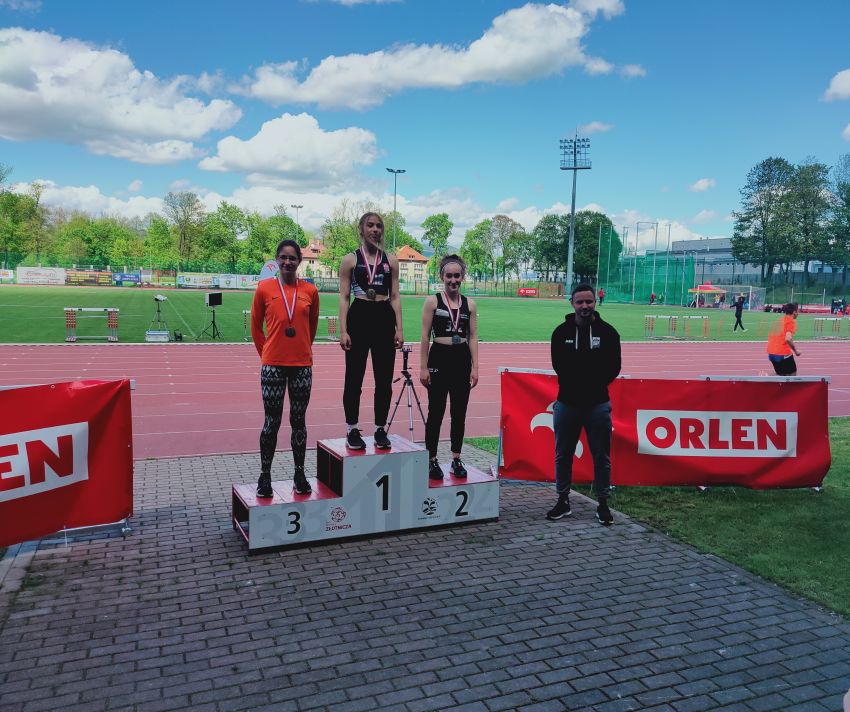 Julia Kruk na drugim miejscu na podium w czarnym stroju z medalem. Za podium widocznia bieżnia i stadion., stadion, domy i drzewa