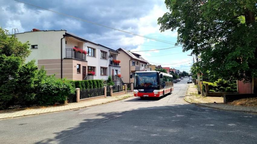 Autobus jedzie ulicą. Po jednej stronie ulicy domy mieszkalne, po drugiej drzewa. Na dalszym planie samochody